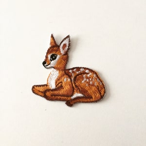 May include: Embroidered patch featuring a fawn in a resting pose. The patch showcases a detailed design with shades of brown, white, and black. The fawn has white spots and is lying down, with its ears perked up.