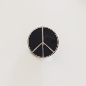 May include: A black peace sign pin with a silver border.