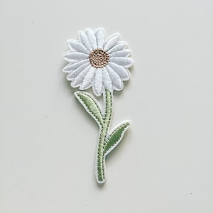 May include: White embroidered daisy flower patch with green leaves and a brown centre.