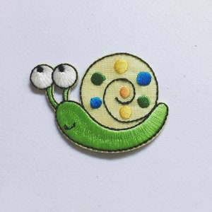 May include: A green snail with white eyes and a yellow shell with colourful dots. The snail is embroidered and has a happy face.