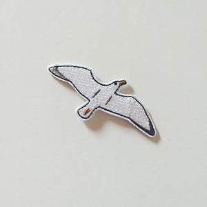 May include: White embroidered seagull patch with black and brown details. The seagull is in flight with its wings spread out.