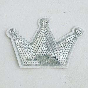 Pode incluir: Um remendo de coroa de lantejoulas prateadas com um suporte de feltro branco.