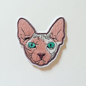 Può includere: Toppa ricamata raffigurante la testa di un gatto Sphynx. La toppa ha un corpo rosa, occhi verdi e un contorno bianco. Il volto del gatto è dettagliato con linee ricamate e la toppa ha un bordo bianco con un contorno marrone.