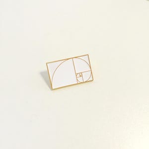 Può includere: Uno spillo in smalto bianco con contorno dorato che rappresenta la spirale di Fibonacci, una sequenza matematica che si trova in natura.