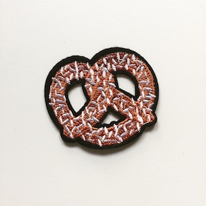 May include: A brown and white embroidered pretzel patch. The pretzel is outlined in black.