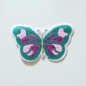 Op de afbeelding: Een groene en roze vlinderapplicatie met pailletten. De vlinder heeft een gouden draadomtrek.