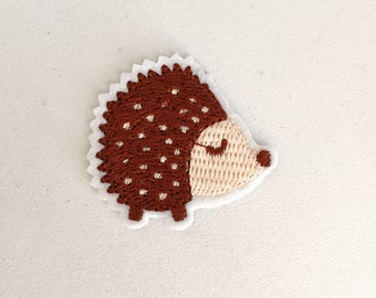 以下が含まれることがあります： 白い縁取りのある、茶色とベージュの刺繍されたハリネズミのパッチ。ハリネズミは、白い点のある茶色の体と、目を閉じたベージュの顔をしています。