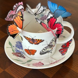 May include: A white teacup and saucer set decorated with butterfly illustrations and floral patterns. Several colorful paper butterflies are arranged around the cup, with a tea bag inside. The set is on a dark wooden surface.