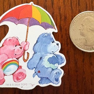 May include: A colorful sticker featuring two Care Bears. One pink bear holds a rainbow ball, the other is blue with a cloud on its belly. They are under a rainbow umbrella. A quarter is next to the sticker. The sticker has the text "TM & © 2024 TCFC".