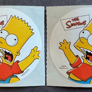 May include: Two round stickers featuring Bart Simpson from The Simpsons cartoon. Bart is yellow with a red shirt and is yelling with his mouth open. The stickers are white with the words "The Simpsons" in a black banner.