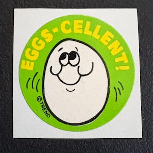 May include: A square sticker with a white background. The sticker features a green circle with the words "EGGS-CELLENT!" in yellow. Inside the circle is a smiling egg character with a black outline. The word "TREND" is printed at the bottom.