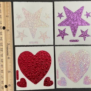 May include: Four sheets of iridescent stickers with a textured finish. Each sheet features a different design: a large star with smaller stars, a large heart with smaller hearts, a purple star with smaller stars, and a white heart with smaller hearts.