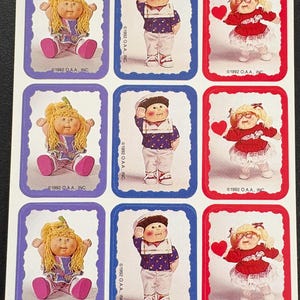 May include: Sheet of vintage Cabbage Patch Kids stickers. The stickers feature various Cabbage Patch Kids dolls in different outfits, including a sailor, a girl in a red dress, and a girl in a patterned dress. Each sticker is framed with a colored border.