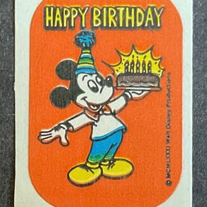May include: A red sticker with a cartoon image of Mickey Mouse wearing a party hat and holding a birthday cake with lit candles. The text "Happy Birthday" is printed in black at the top of the sticker.