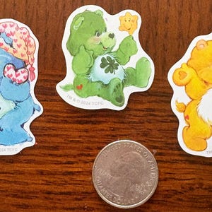 May include: Three Care Bear stickers featuring a blue bear in a nightcap, a green bear with a four-leaf clover, and a yellow bear with a sun. A U.S. quarter is included for size comparison. The stickers are on a brown wooden surface.
