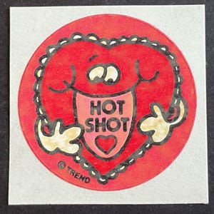 May include: Red and white sticker with a cartoon heart character with a tongue sticking out and the words "HOT SHOT" on it. The heart character is wearing a small heart on its chest. The sticker has a black border and the copyright symbol and "TREND" are printed at the bottom.