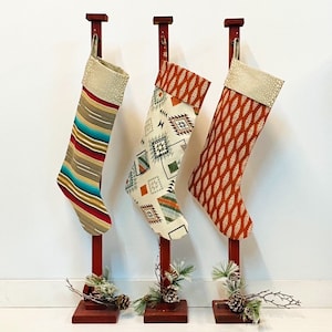May include: Three red wooden stocking holders with three different patterned Christmas stockings. The stockings are hung on the holders and have a cream-colored cuff. The stockings are decorated with geometric patterns in shades of orange, brown, and blue.
