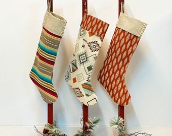 May include: Three red wooden stocking holders with three different patterned Christmas stockings. The stockings are hung on the holders and have a cream-colored cuff. The stockings are decorated with geometric patterns in shades of orange, brown, and blue.