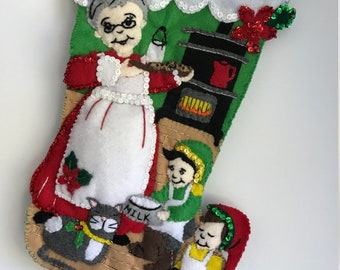 May include: A felt Christmas stocking with a green background and a white, fluffy cuff. The stocking features a woman with grey hair baking cookies in a kitchen with a cat and two children. The stocking is decorated with sequins and beads.