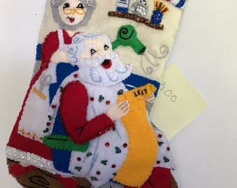 May include: A felt Christmas stocking featuring Santa Claus and a woman in a red dress. Santa is seated in a barber's chair receiving a haircut. The woman is holding a pair of scissors. The stocking is adorned with sequins and beads.