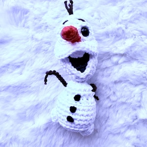 May include: A handmade crochet snowman with a large open mouth, black eyes, and a red nose. The snowman has three black buttons and brown twig arms and hair. The snowman is made of white yarn and is set against a fluffy white background.