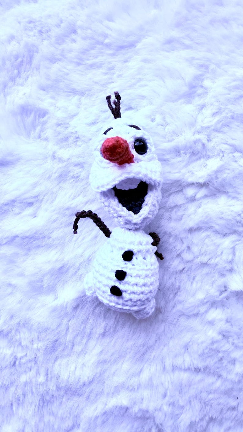May include: A handmade crochet snowman with a large open mouth, black eyes, and a red nose. The snowman has three black buttons and brown twig arms and hair. The snowman is made of white yarn and is set against a fluffy white background.
