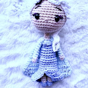 May include: A handmade crochet doll with a light skin tone, black eyes, and purple eyelashes. The doll has white hair and wears a blue dress and cape. The doll is lying on a white, fluffy surface.