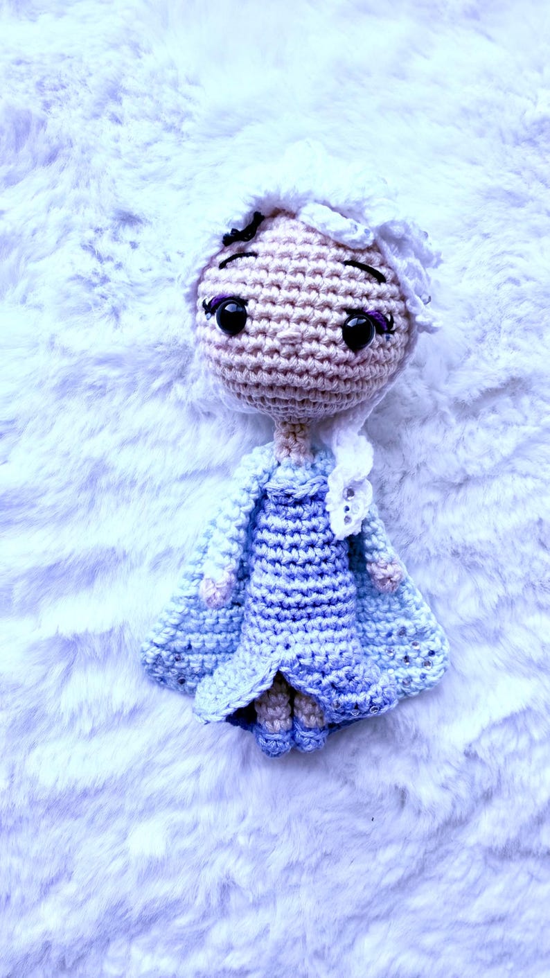 May include: A handmade crochet doll with a light skin tone, black eyes, and purple eyelashes. The doll has white hair and wears a blue dress and cape. The doll is lying on a white, fluffy surface.