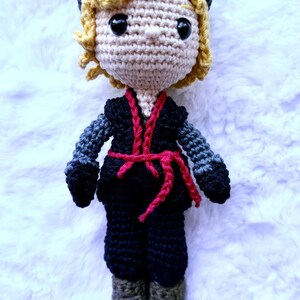May include: A handmade crochet doll featuring a light skin tone, blonde curly hair, and a dark blue hat. The doll wears a black outfit with gray sleeves and a red belt. The doll is approximately 8 inches tall.