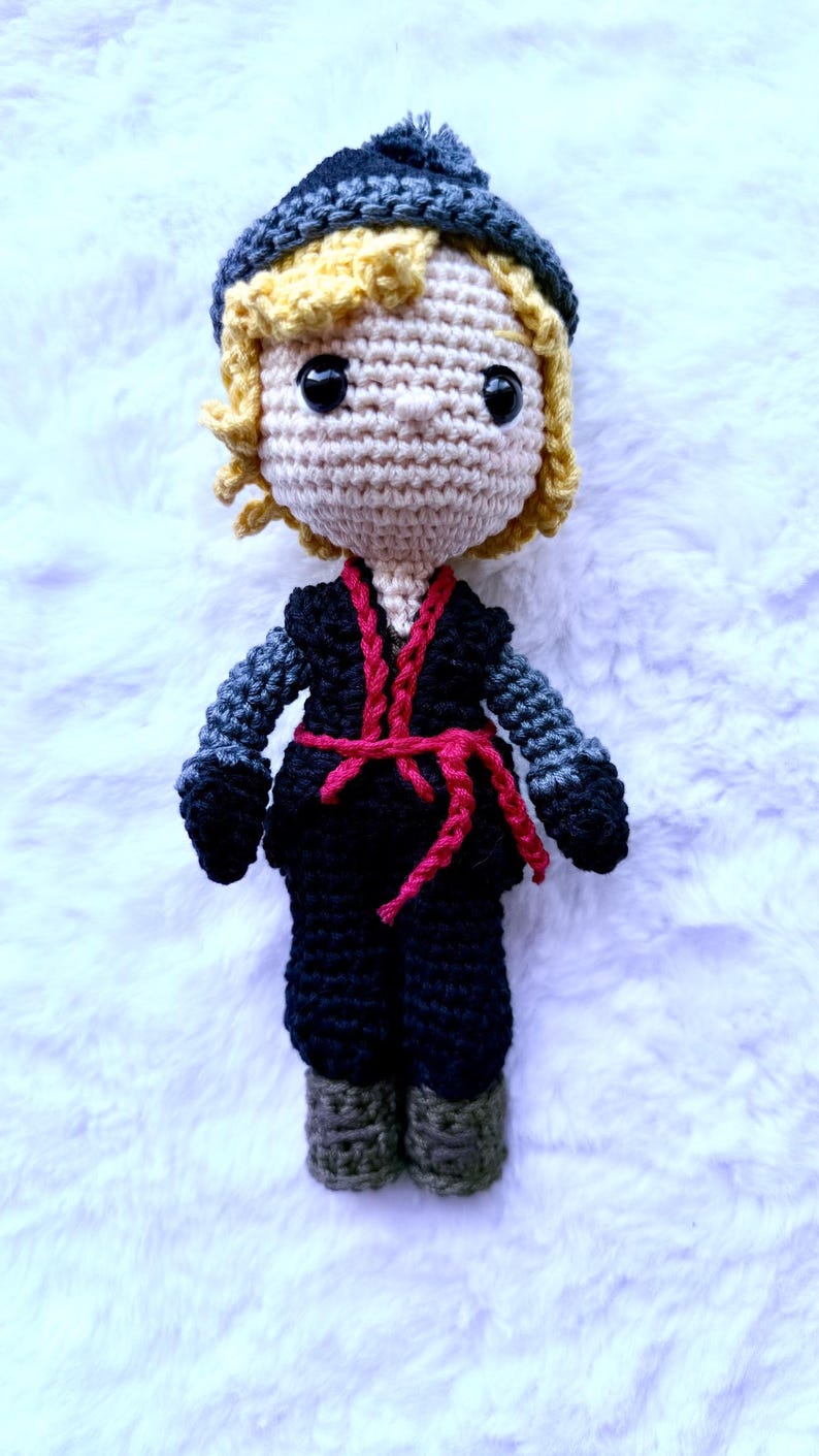 May include: A handmade crochet doll featuring a light skin tone, blonde curly hair, and a dark blue hat. The doll wears a black outfit with gray sleeves and a red belt. The doll is approximately 8 inches tall.