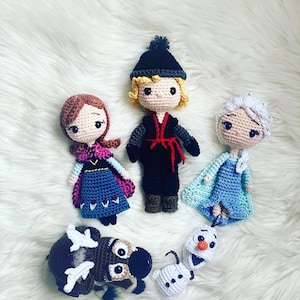 May include: Crocheted dolls of characters from the animated film Frozen, including Anna, Kristoff, Elsa, Olaf, and Sven. The dolls are handmade with yarn and feature detailed facial expressions and clothing. The dolls are arranged on a white, fluffy surface.
