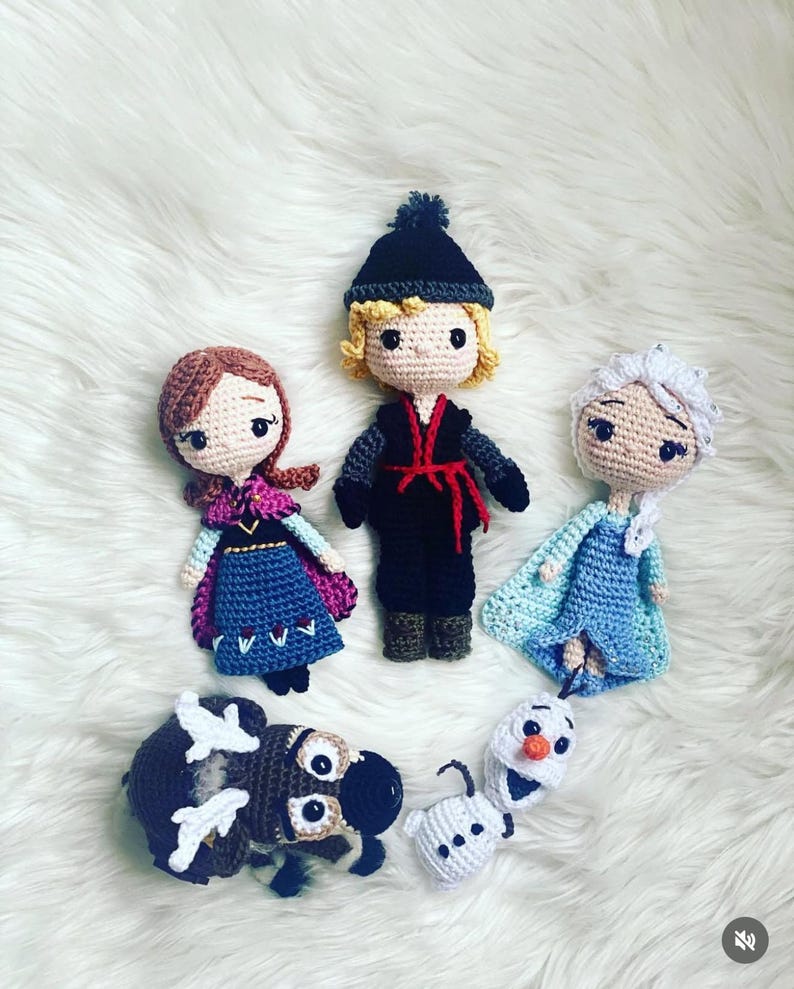 May include: Crocheted dolls of characters from the animated film Frozen, including Anna, Kristoff, Elsa, Olaf, and Sven. The dolls are handmade with yarn and feature detailed facial expressions and clothing. The dolls are arranged on a white, fluffy surface.