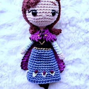 May include: Crocheted doll featuring a character with auburn hair, wearing a purple cape, blue skirt, and black shoes. The doll has a light skin tone and large black eyes. The doll is handmade and sits on a white, fluffy surface.