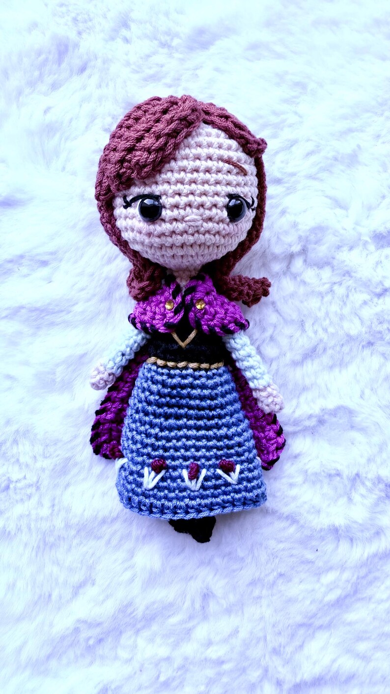 May include: Crocheted doll featuring a character with auburn hair, wearing a purple cape, blue skirt, and black shoes. The doll has a light skin tone and large black eyes. The doll is handmade and sits on a white, fluffy surface.