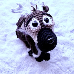 May include: A handmade crochet reindeer toy with large black eyes, a black nose, and brown and white fur. The reindeer has small ears and antlers. The toy is lying on a white, fluffy surface.