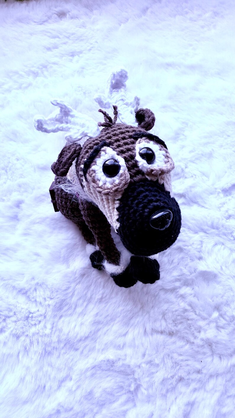 May include: A handmade crochet reindeer toy with large black eyes, a black nose, and brown and white fur. The reindeer has small ears and antlers. The toy is lying on a white, fluffy surface.