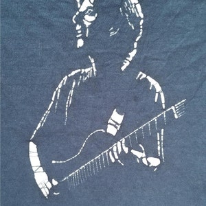 May include: Navy blue t-shirt featuring a white graphic of a musician playing a guitar. The design is a stylized silhouette, with the musician's form and instrument clearly defined against the dark fabric. The shirt is made of a soft material.