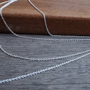 May include: Close-up of multiple strands of delicate silver chain necklaces. The chains are displayed on a wooden surface, showcasing their intricate links and reflective quality. The chains are layered, creating a sense of depth.
