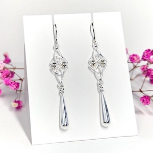 Könnte beinhalten: Ein Paar silberfarbene Ohrringe mit keltischen Knoten. Jeder Ohrring hat ein detailliertes Knotendesign mit kleinen Perlenakzenten, das an einem Haken befestigt ist. Die Ohrringe haben einen tropfenförmigen Anhänger. Der Hintergrund ist weiß mit rosa Blumen.