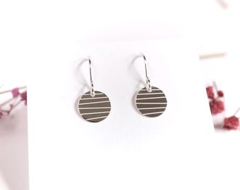 May include: A pair of silver drop earrings. Each earring features a circular disc with horizontal lines. The earrings hang from a simple hook. The background is white, with pink flowers on the left and right.