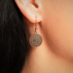K&ouml;nnte beinhalten: Kupferfarbener Ohrring mit einem kreisf&ouml;rmigen Anh&auml;nger mit einem aufwendigen, detaillierten Design. Der Ohrring h&auml;ngt an einem einfachen Haken und zeigt einen Vintage- oder Boh&egrave;me-Stil. Das Design ist auf dem Anh&auml;nger zentriert.