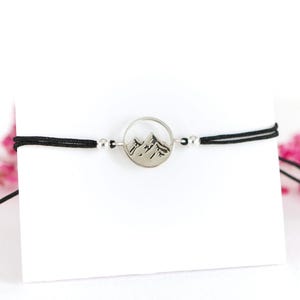 May include: A black cord bracelet featuring a circular silver-toned charm with a mountain range cutout design. The charm is flanked by two small silver-toned beads on either side, connecting the cord to the central mountain motif.