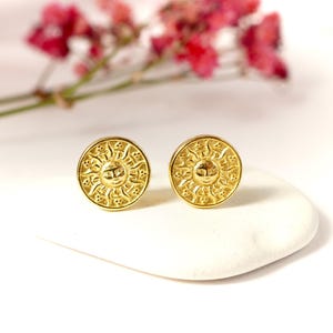 May include: A pair of gold-toned earrings with a sun design. The earrings are round and have a detailed sun face with rays.
