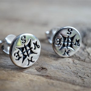 Puede incluir: Un par de pendientes de botón plateados. Cada pendiente presenta un diseño de brújula con los puntos cardinales, Norte, Sur, Este y Oeste, marcados en la cara. Los pendientes están sobre una superficie de madera.