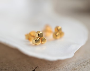 May include: Gold-colored stud earrings with a geometric honeycomb design. Each earring features three connected hexagonal shapes. The earrings are displayed on a white, petal-shaped surface, with a blurred background.