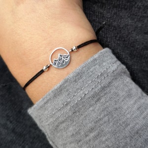 Puede incluir: Una pulsera con un dije de montaña plateado con un cordón negro. El dije presenta una cordillera dentro de un marco circular. Pequeñas cuentas plateadas acentúan el cordón. La pulsera se muestra en una muñeca.
