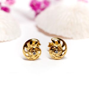 Puede incluir: Un par de pendientes dorados. Cada pendiente presenta un diseño en espiral con una pequeña gema transparente en el centro. Los pendientes están colocados sobre un fondo blanco con acentos borrosos en rosa y blanco.