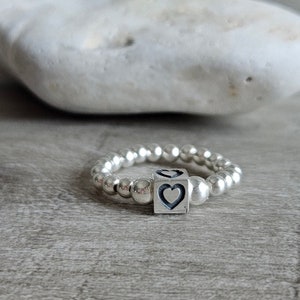Peut inclure: Une bague en argent avec un petit charme en forme de cube orné de deux cœurs. La bague est composée de petites perles en argent.