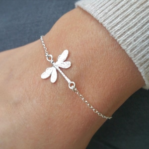 Könnte beinhalten: Ein silbernes Kettenarmband mit einem zarten Libellen-Anhänger. Die Libelle hat detaillierte Flügel und eine strukturierte Oberfläche.