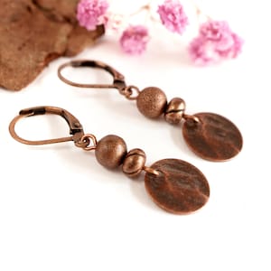Puede incluir: Un par de pendientes colgantes de color cobre. Cada pendiente presenta un cierre de palanca, una cuenta redonda grande, una cuenta más pequeña y un disco martillado. Flores rosas y un trozo de corteza en el fondo.